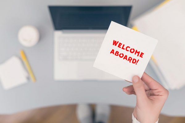 Photo of a Caucasian individual's hand; they're holding a small white note that says "Welcome Aboard!" in red colored text.